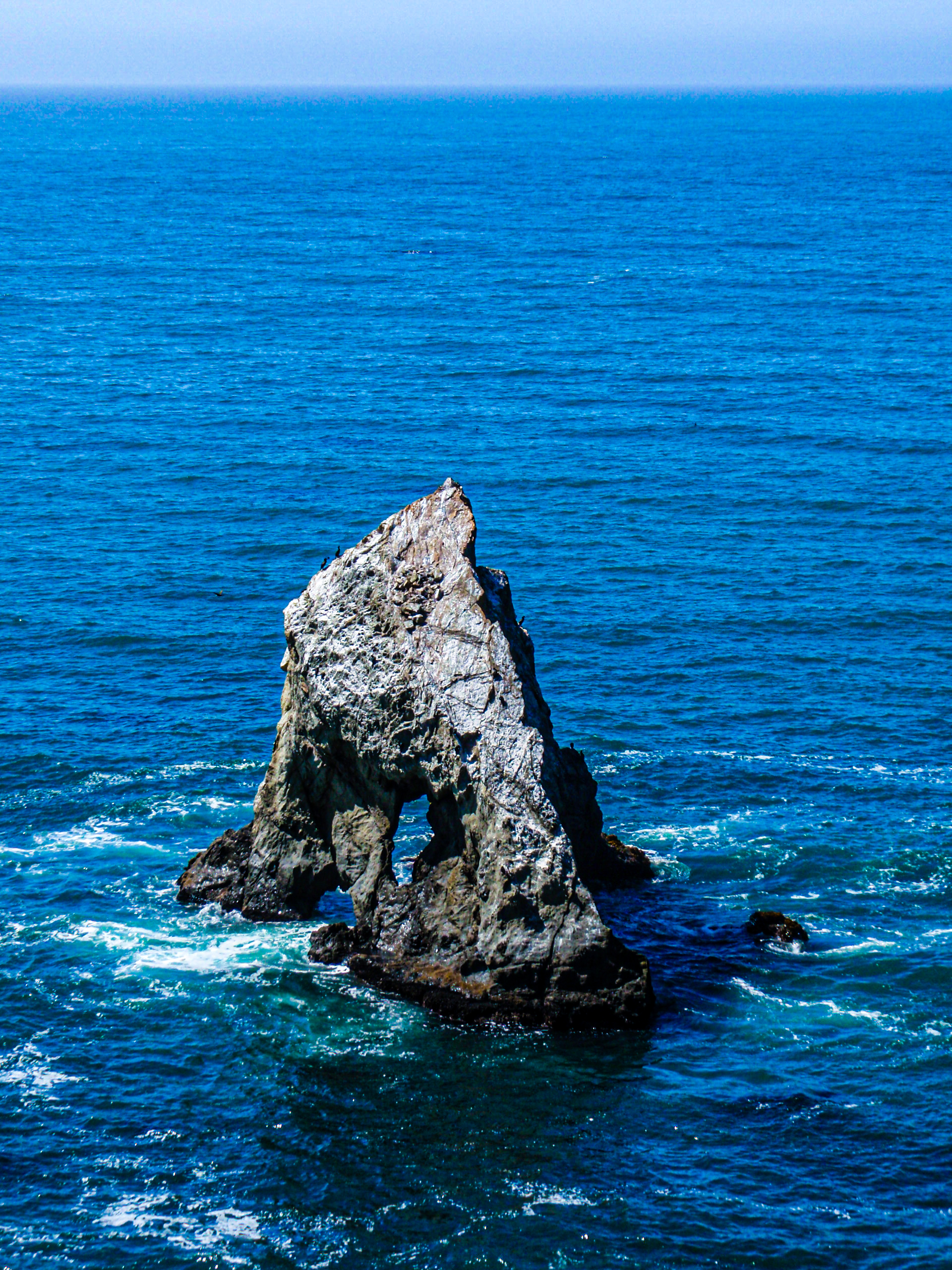 A arched rock protruding from the ocean with deep blue water and grey, intricate stone.