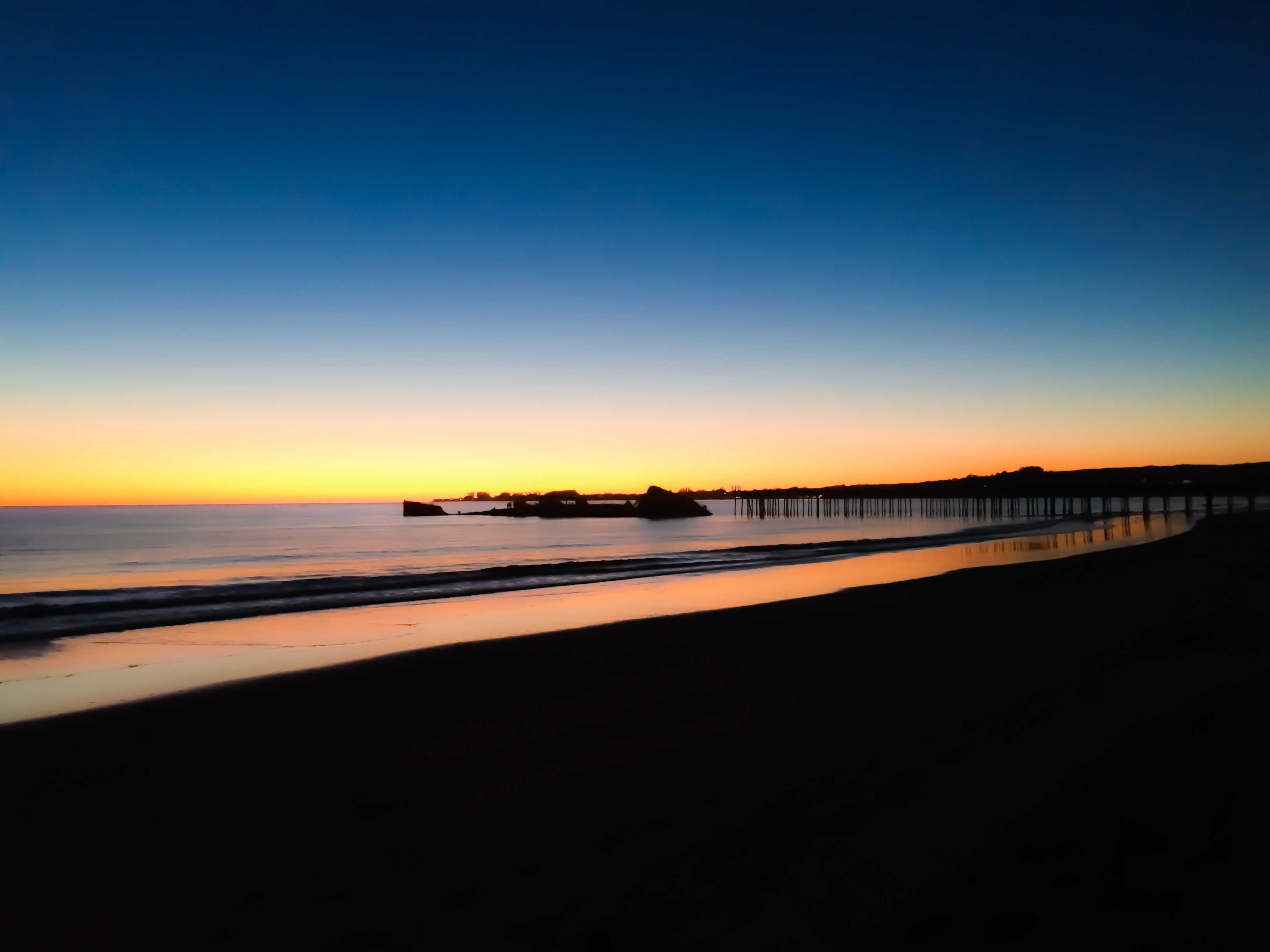 The coastline after sunset with the remains of an old pier extending in front of the pink horizon.