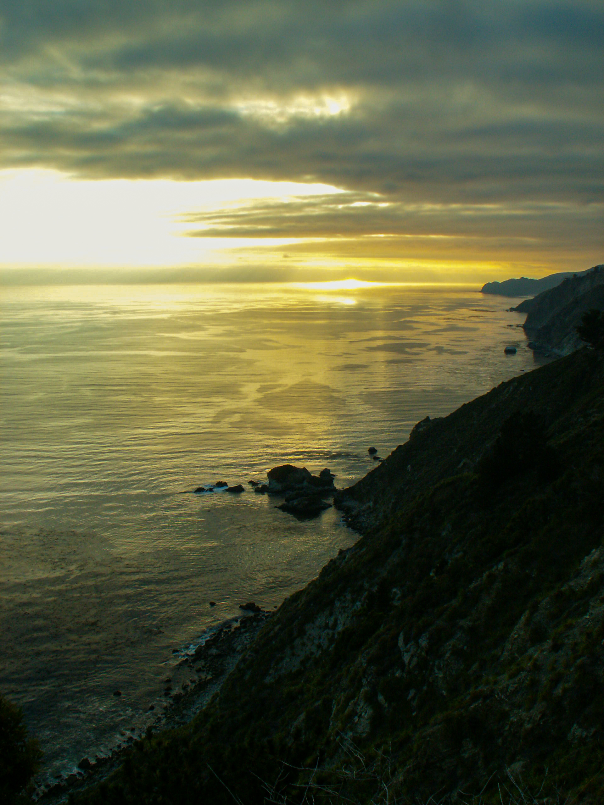 A stretch of coastline extending to the sunset-orange horizon with textured ocean rippling west.